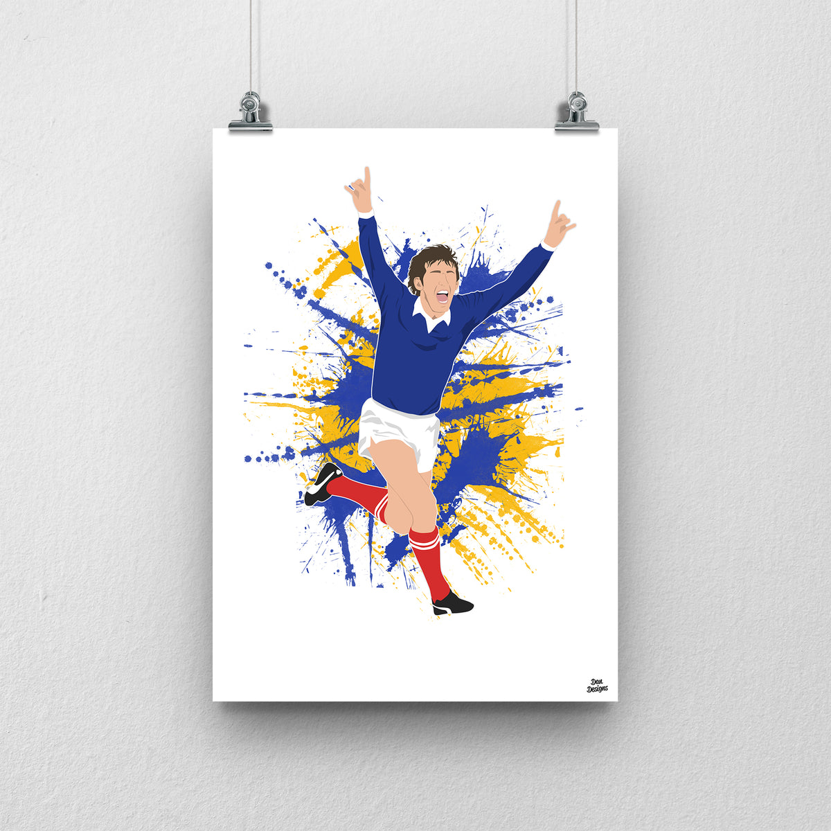 Kenny Dalglish celebrating with both arms raised in Scotland’s navy shirt, white shorts and red socks, illustrated with blue and yellow paint splashes on white.