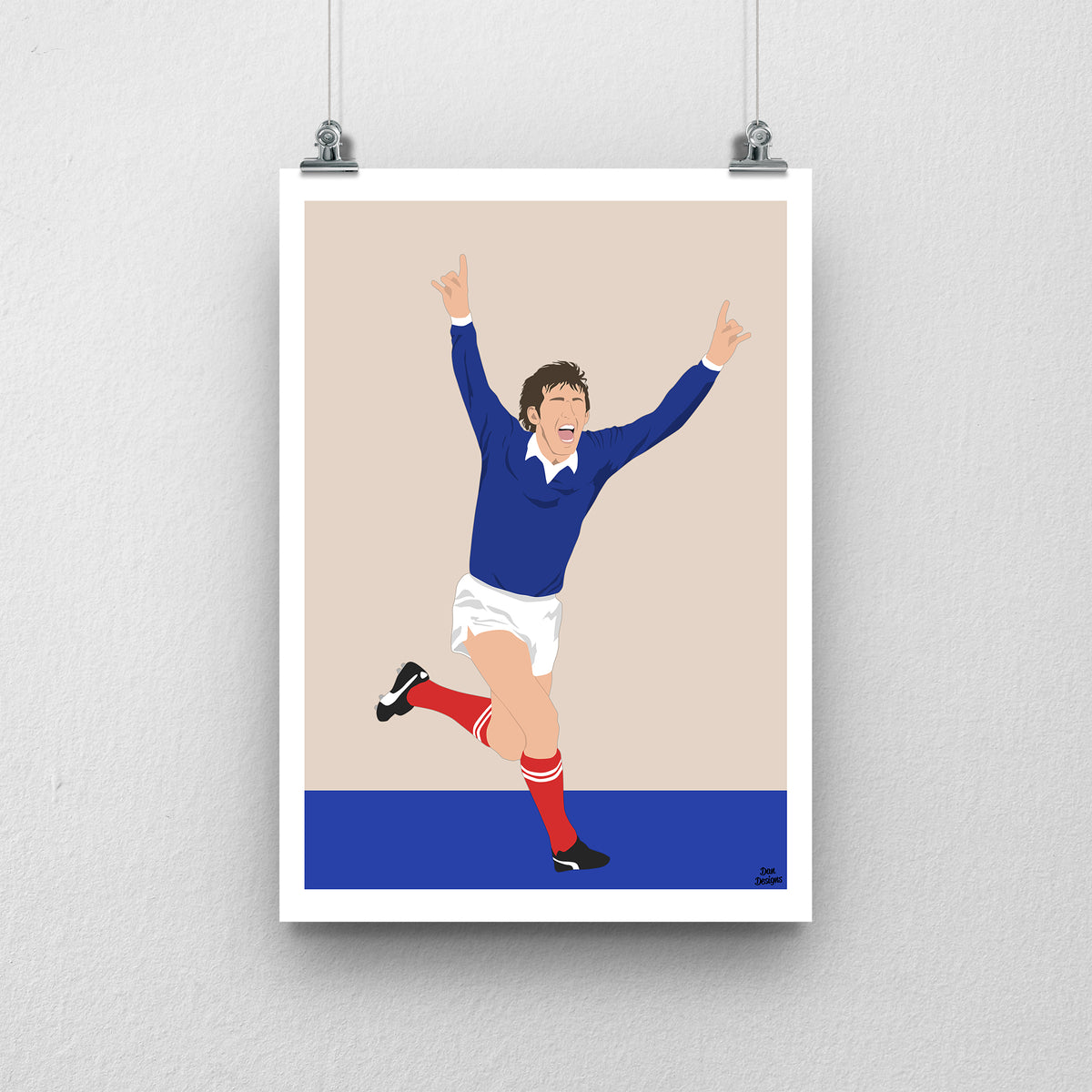 Kenny Dalglish celebrating with both arms raised in Scotland’s navy shirt, white shorts and red socks, illustrated with a beige background, blue base and white border.